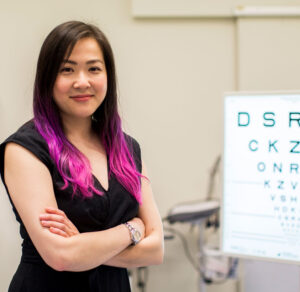 Photograph of Dr Flora Hui standing face to camera in a consultation room with an eye chart to her left hand-side.