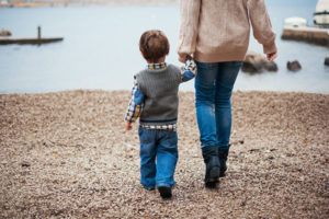 Photo of a mother and her son shown from the back, holding hands and walking towards the sea